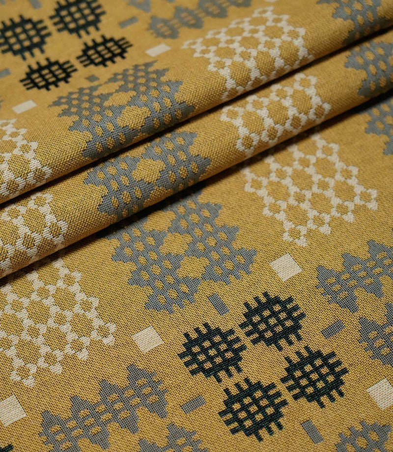 Kinfolk fabric arranged in soft folds, displaying repeating Scandi-style geometric motifs in charcoal, grey and cream against a mustard yellow ground.