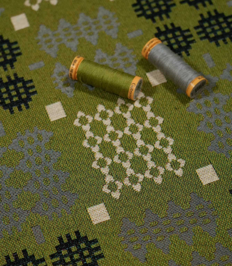 Kinfolk geometric fabric with green and grey thread spools resting on the surface, highlighting the olive green ground, woven texture and contrasting motifs.