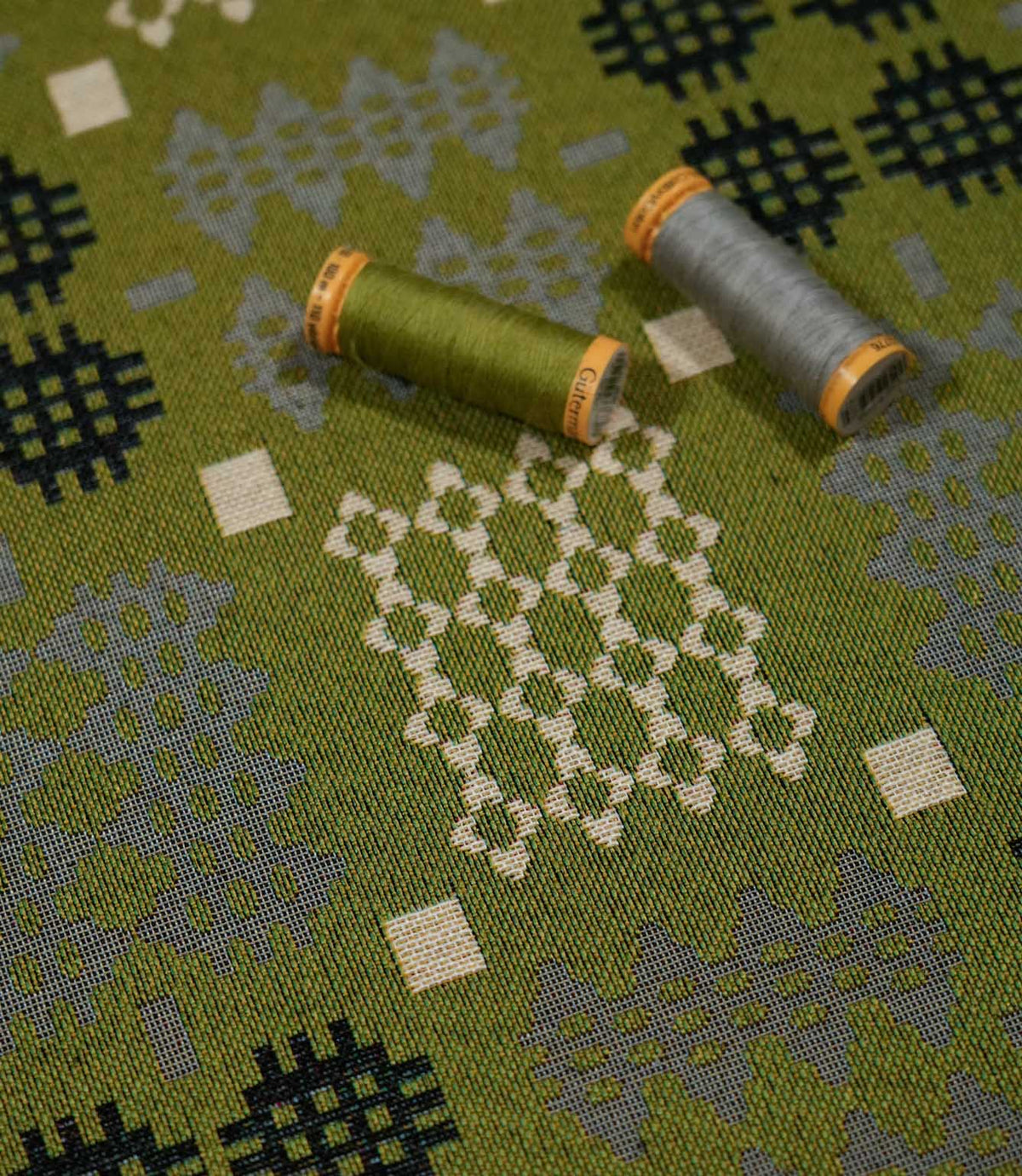 Kinfolk geometric fabric with green and grey thread spools resting on the surface, highlighting the olive green ground, woven texture and contrasting motifs.