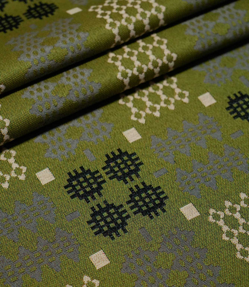 Kinfolk fabric arranged in neat folds, displaying repeating Scandi-style geometric motifs in black, grey and cream against a textured olive green background.