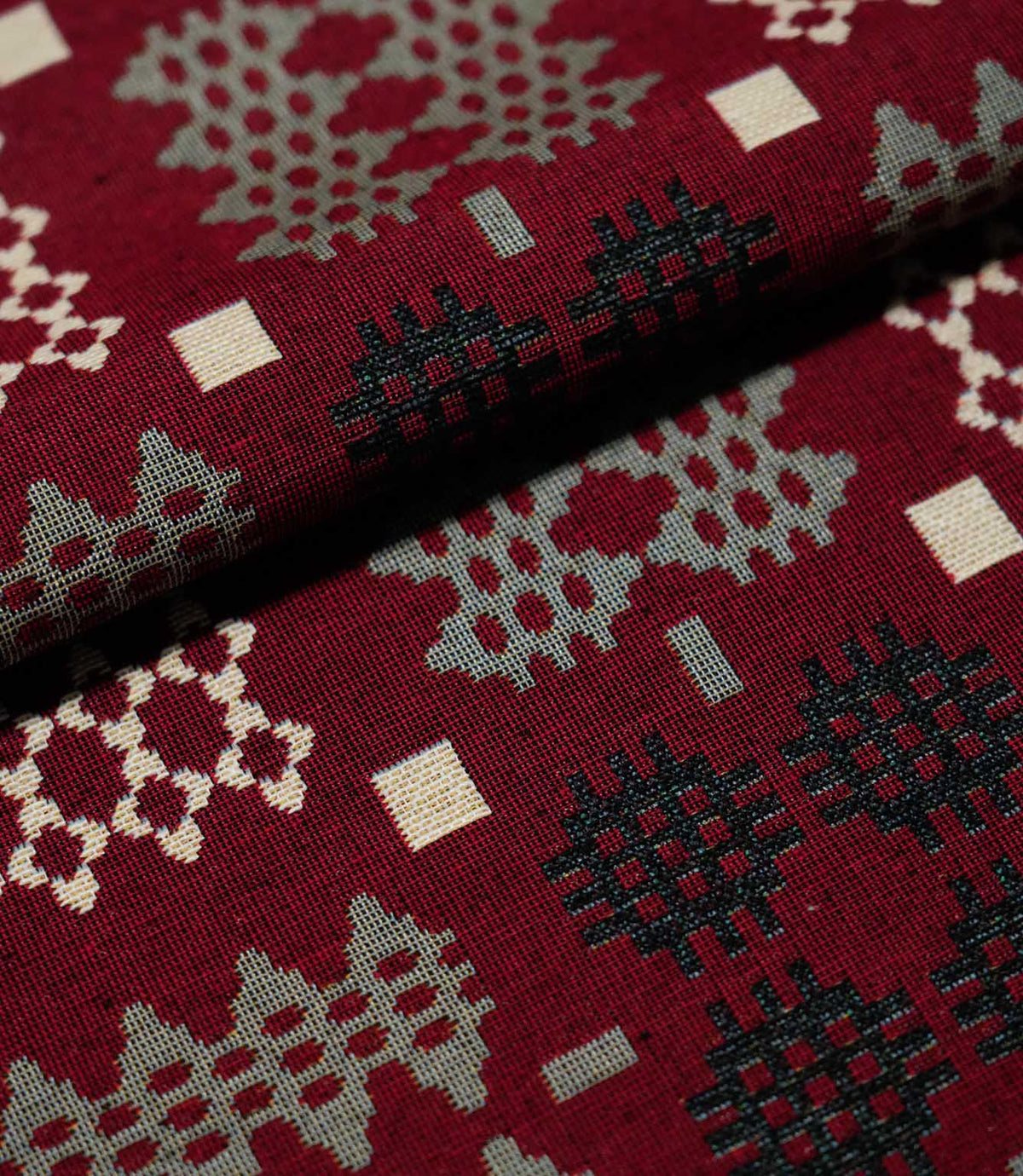 Close-up of folded Kinfolk fabric, showing the scale and woven detail of its black, grey and cream geometric motifs on a rich red ground.