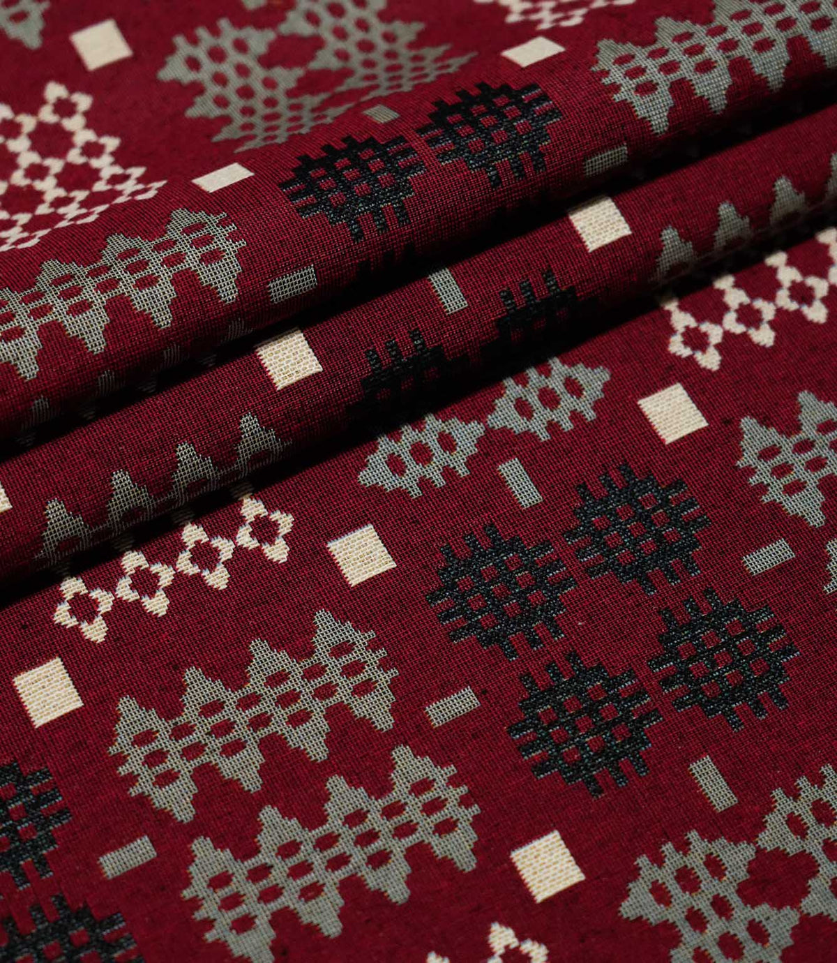 Kinfolk fabric arranged in neat folds, displaying repeating geometric motifs in black, soft grey and cream against a textured deep red background.