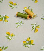 Lemon Grove Natural citrus print fabric photographed with green and yellow sewing thread spools for scale, highlighting the small-scale lemon motifs, white blossoms and natural woven texture.
