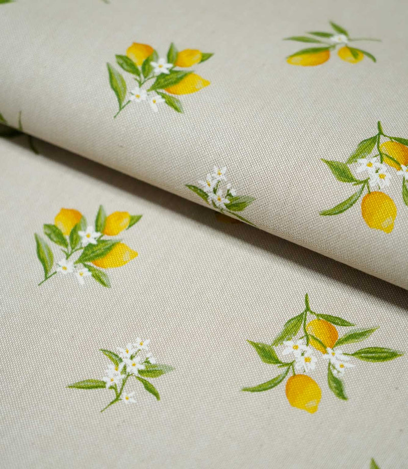 Lemon Grove Natural fabric folded over itself, showing the scattered yellow lemon print, fresh green leaves and delicate white citrus flowers on a natural beige background.