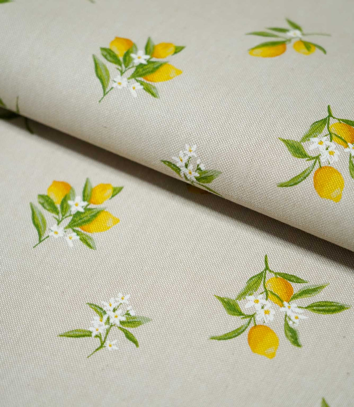 Lemon Grove Natural fabric folded over itself, showing the scattered yellow lemon print, fresh green leaves and delicate white citrus flowers on a natural beige background.