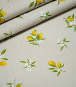 Lemon Grove Natural botanical fabric arranged in overlapping folds, displaying its repeating yellow lemon branches, green foliage and white blossoms across a textured woven surface.