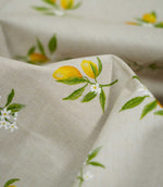 Detailed close-up of Lemon Grove Natural fabric showing yellow lemons, green leaves and tiny white blossoms printed onto a textured beige woven surface, with soft fabric folds in the background.