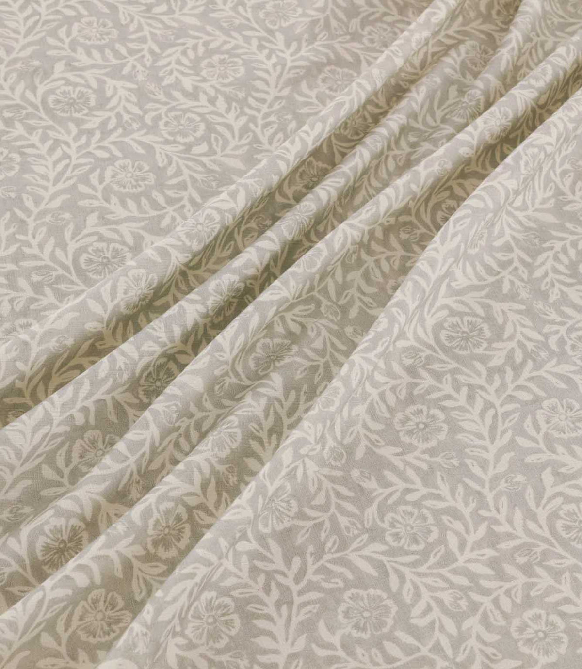Manorville Gull floral fabric arranged in soft, flowing folds, showing its delicate ivory flowers, intertwining leafy vines and warm grey-beige background, with the botanical pattern continuing naturally across the draped material.
