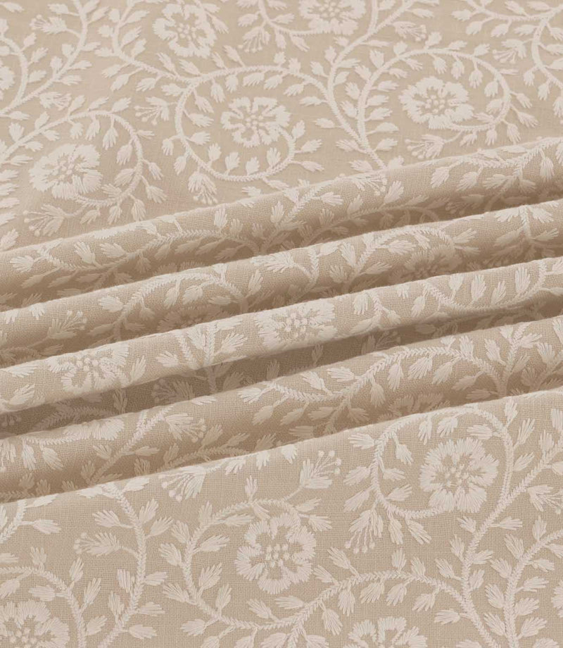 Close-up of Marion Quaker fabric draped in soft folds, highlighting the intricate ivory embroidered flowers, trailing vines and delicate foliage on a natural beige woven background.