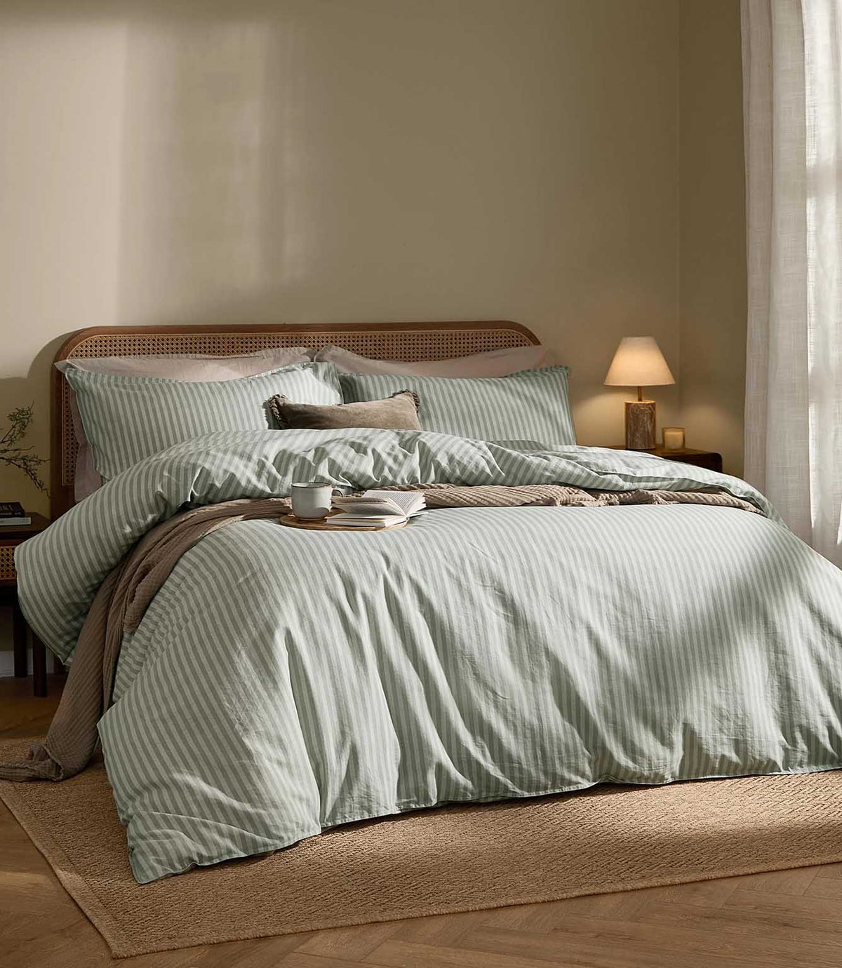 Marlow Stripe Duvet Cover Set in Artichoke/Sage, styled on a double bed with soft sage green and off-white stripes, neutral cushions, a taupe throw and cane headboard.