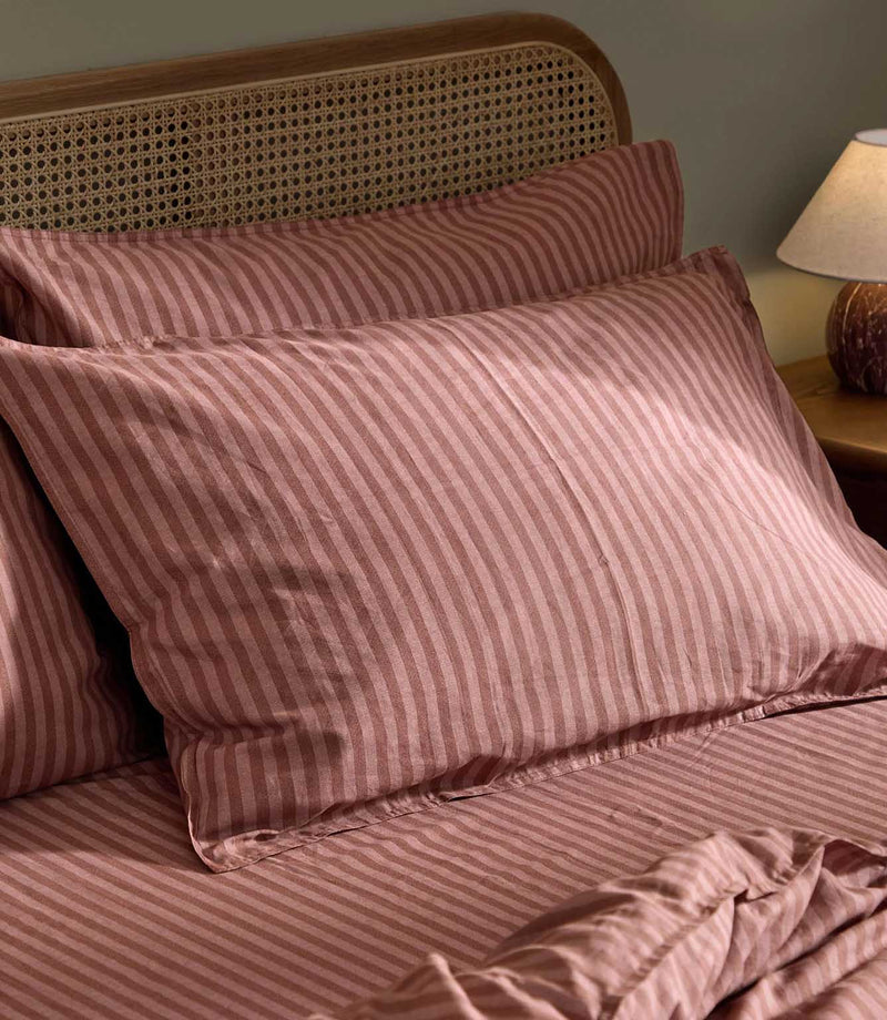 Close-up of Marlow Stripe pillowcases in Chocolate/Rosewood showing alternating rose pink and chocolate brown stripes, with matching bedding and a natural cane headboard.