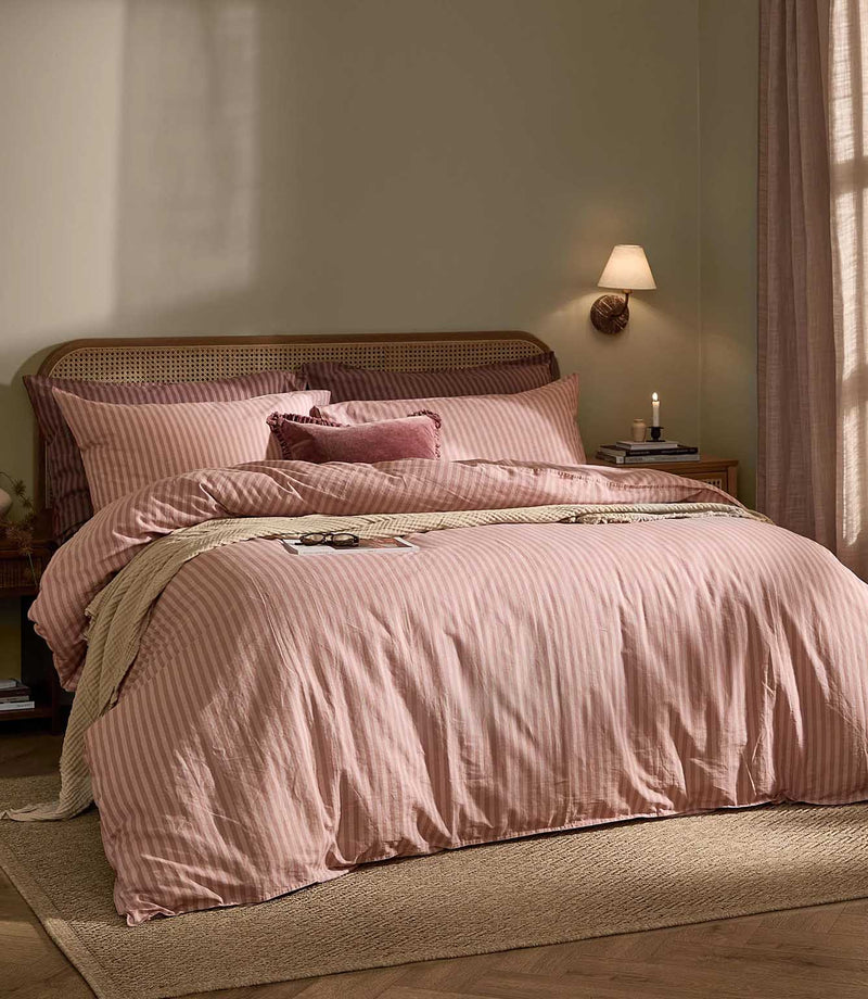 Marlow Stripe duvet cover set in soft blush pink and rose stripes, styled on a bed with coordinating pillows, a textured throw and natural cane headboard.