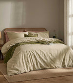 Marlow Stripe duvet cover set in soft olive green and cream, styled on a bed with coordinating striped pillowcases, green cushions, a textured throw and cane headboard.