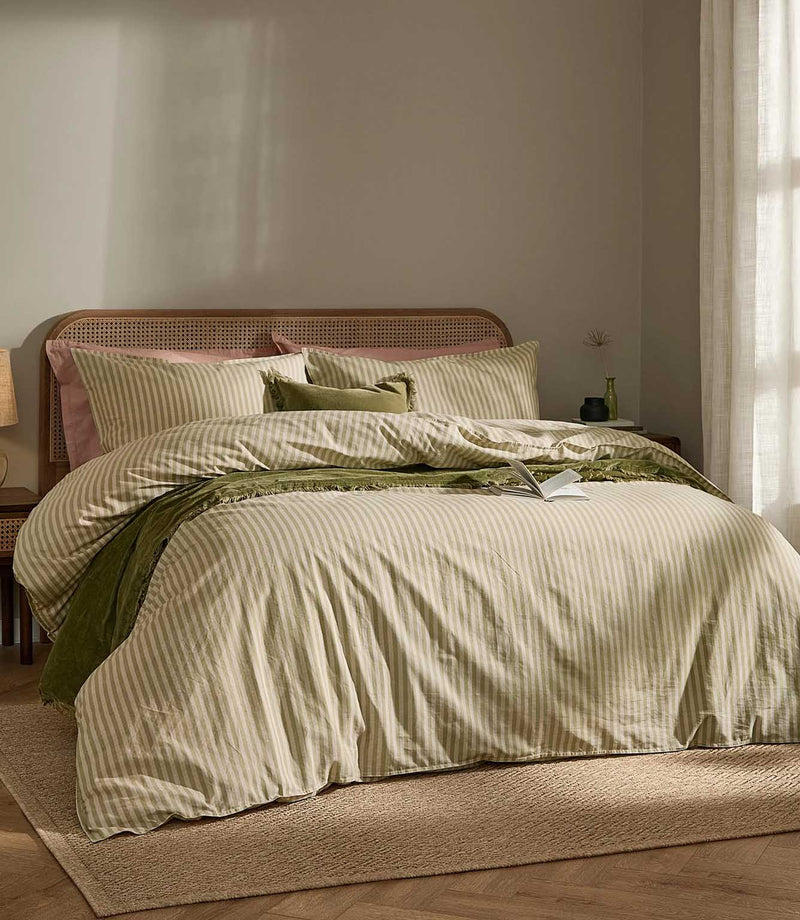 Marlow Stripe duvet cover set in soft olive green and cream, styled on a bed with coordinating striped pillowcases, green cushions, a textured throw and cane headboard.