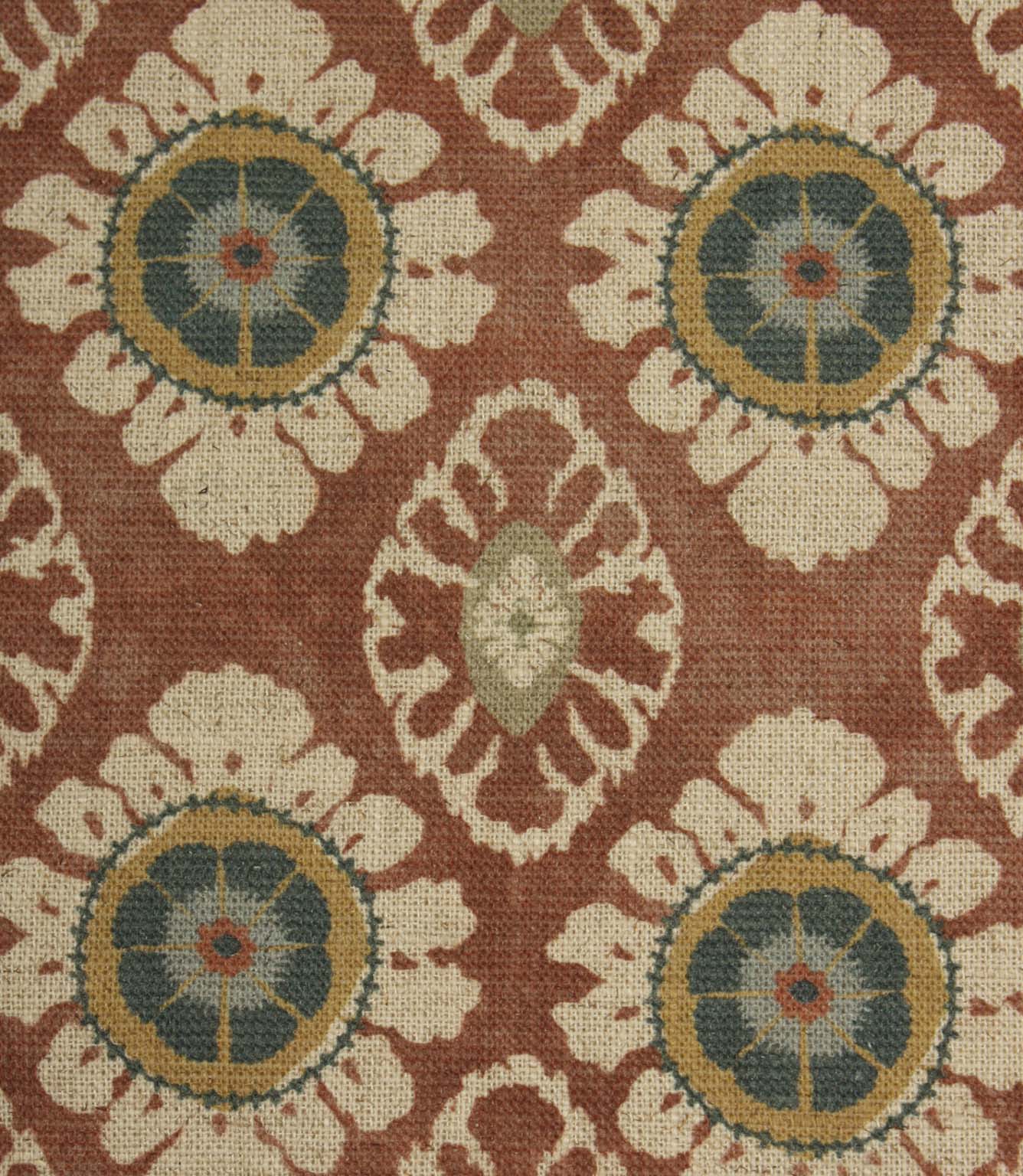 Close-up of terracotta red decorative fabric with stylised floral medallions and intricate cream patterning on a textured weave.