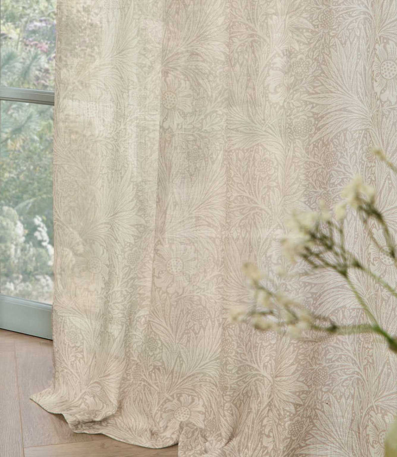 Close-up of Morris & Co Marigold sheer curtain fabric in soft cream and linen beige, showing the delicate floral and foliage pattern filtering natural light beside a window.