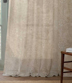 Full-length Morris & Co Marigold sheer curtain in cream and linen beige, softly draping onto a wooden floor beside a pale green framed window.