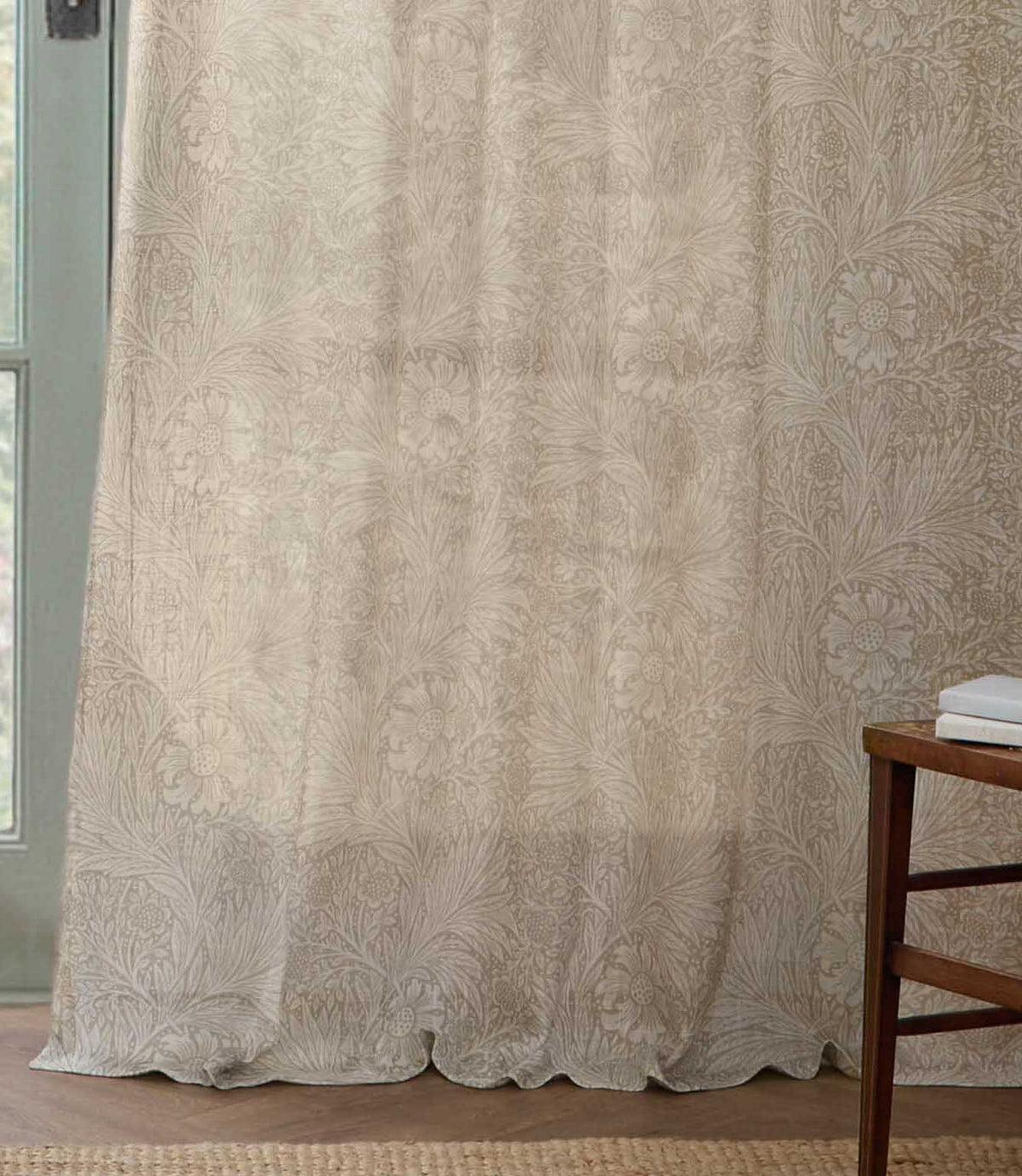 Full-length Morris & Co Marigold sheer curtain in cream and linen beige, softly draping onto a wooden floor beside a pale green framed window.
