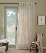 Morris & Co Marigold sheer curtain in cream and linen beige hanging beside a pale green glazed door, styled in a relaxed room with natural wood furniture and woven accessories.