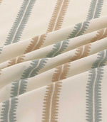 Close-up of Meschutt Saltwater embroidered stripe fabric in soft folds, showing raised muted blue-grey and warm tan zigzag stripes with fine central stitching on ivory fabric.