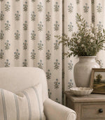 Novi Ink botanical print curtains with delicate blue flowers and green stems on a natural cream background, styled beside a neutral armchair and wooden side table with a ceramic vase of flowers.