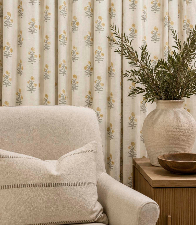Novi Turmeric botanical print curtains featuring small mustard yellow flowers and soft grey-green stems on a natural cream background, styled with a neutral armchair and wooden side table.