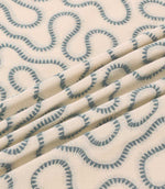 Close-up of Orient Saltwater fabric draped in folds, featuring muted blue embroidered squiggle patterns on a soft cream woven background.