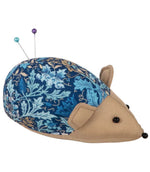 Hedgehog pincushion with a navy blue, turquoise and gold woodblock floral patterned back, beige fabric face and ears, with two sewing pins in the padded cushion.