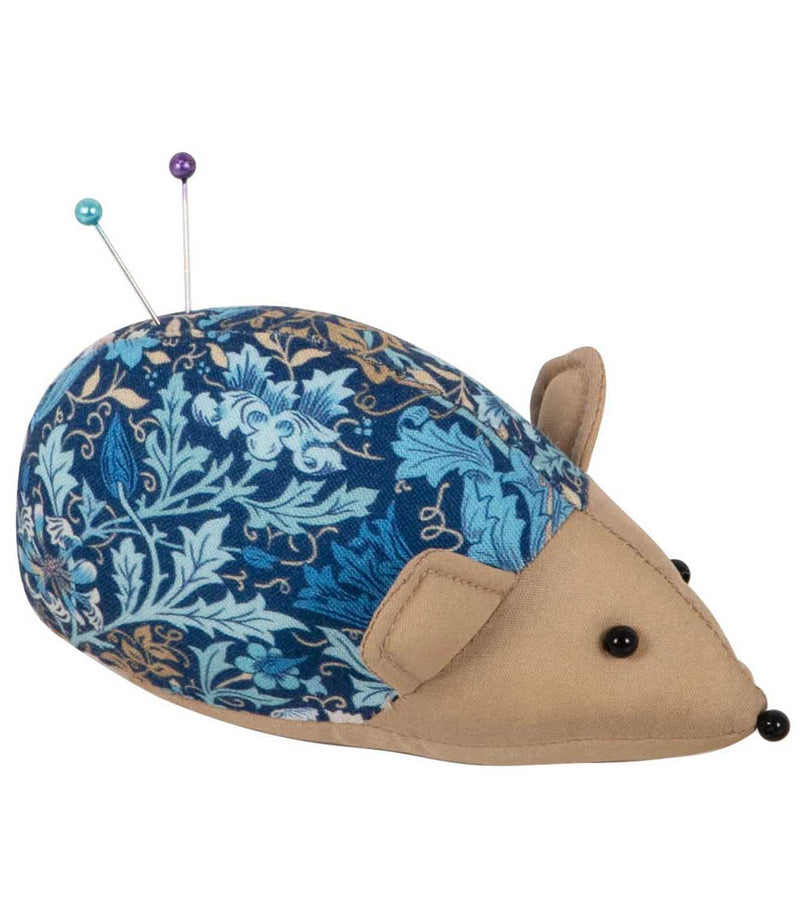 Hedgehog pincushion with a navy blue, turquoise and gold woodblock floral patterned back, beige fabric face and ears, with two sewing pins in the padded cushion.