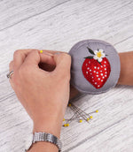 Grey wrist pincushion worn on the wrist while sewing, featuring an embroidered red strawberry with white flower and green leaves, with dressmaking pins nearby.