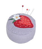 Grey fabric wrist pincushion with embroidered red strawberry, white flower and green leaves, shown holding four colourful sewing pins
