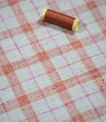Painswick Check Russett fabric laid flat with a coordinating russet orange thread spool, showcasing the repeating orange and pink check pattern, natural cream background and textured woven finish.