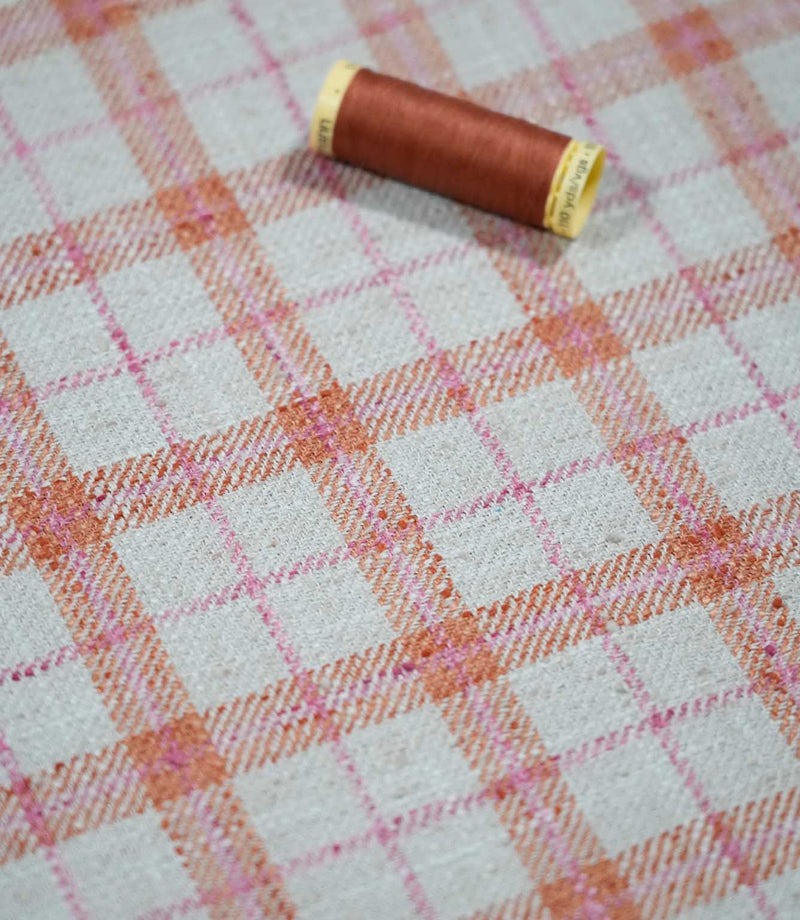 Painswick Check Russett fabric laid flat with a coordinating russet orange thread spool, showcasing the repeating orange and pink check pattern, natural cream background and textured woven finish.