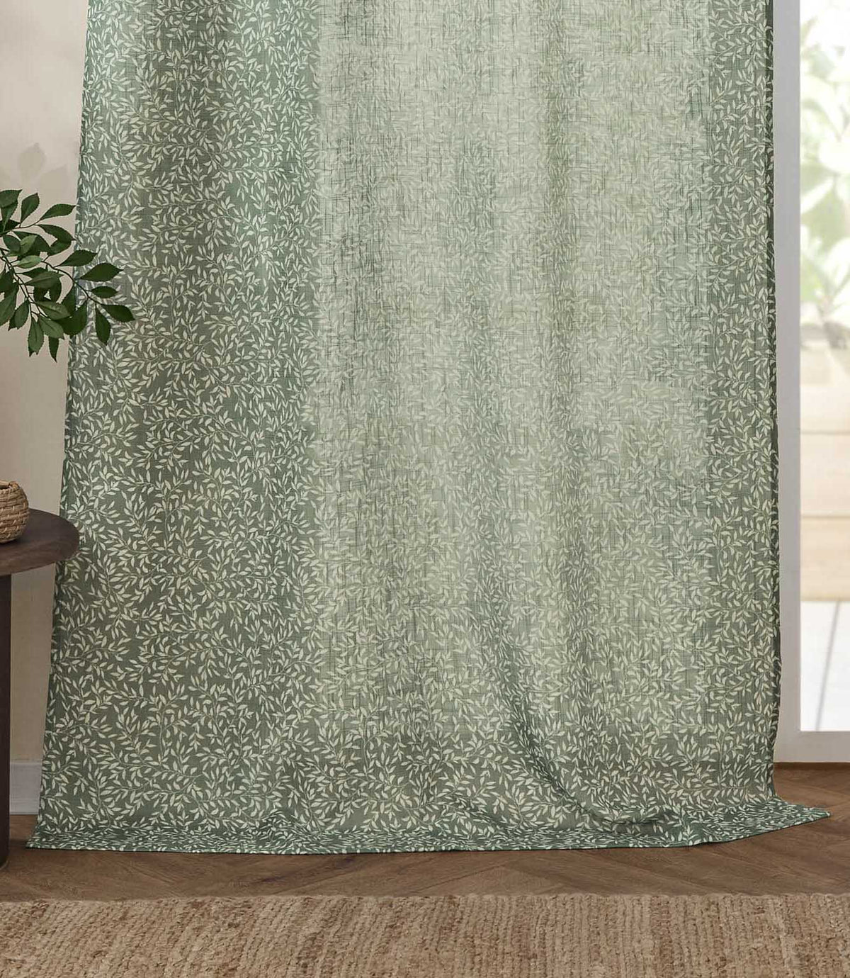 Full-length Perrie Leaf sheer curtain panel in forest green and soft white, softly filtering daylight beside a glazed door in a natural living space.