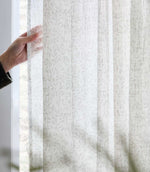 Close-up of natural cream Perrie Leaf sheer curtain fabric held against a bright window, showing its lightweight, semi-transparent woven texture and subtle beige leaf pattern.
