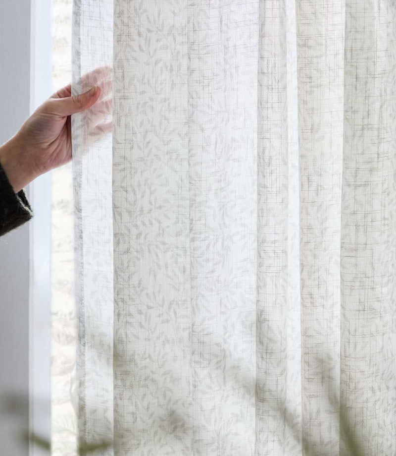 Close-up of natural cream Perrie Leaf sheer curtain fabric held against a bright window, showing its lightweight, semi-transparent woven texture and subtle beige leaf pattern.