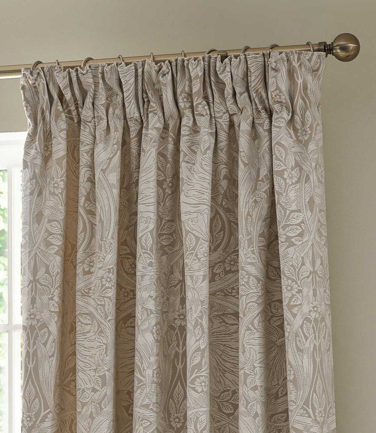 Morris & Co Pimpernel cream and beige jacquard pencil pleat curtain hanging from a metal curtain pole, showing the detailed woven botanical pattern and gathered heading.