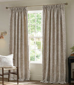 Pair of full-length Morris & Co Pimpernel jacquard curtains in cream and beige framing a bright window, styled with natural wood furniture in a neutral living room.