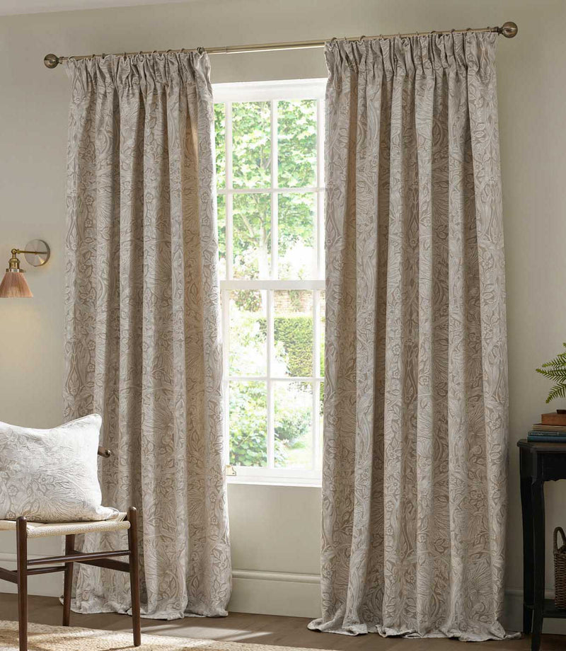 Pair of full-length Morris & Co Pimpernel jacquard curtains in cream and beige framing a bright window, styled with natural wood furniture in a neutral living room.
