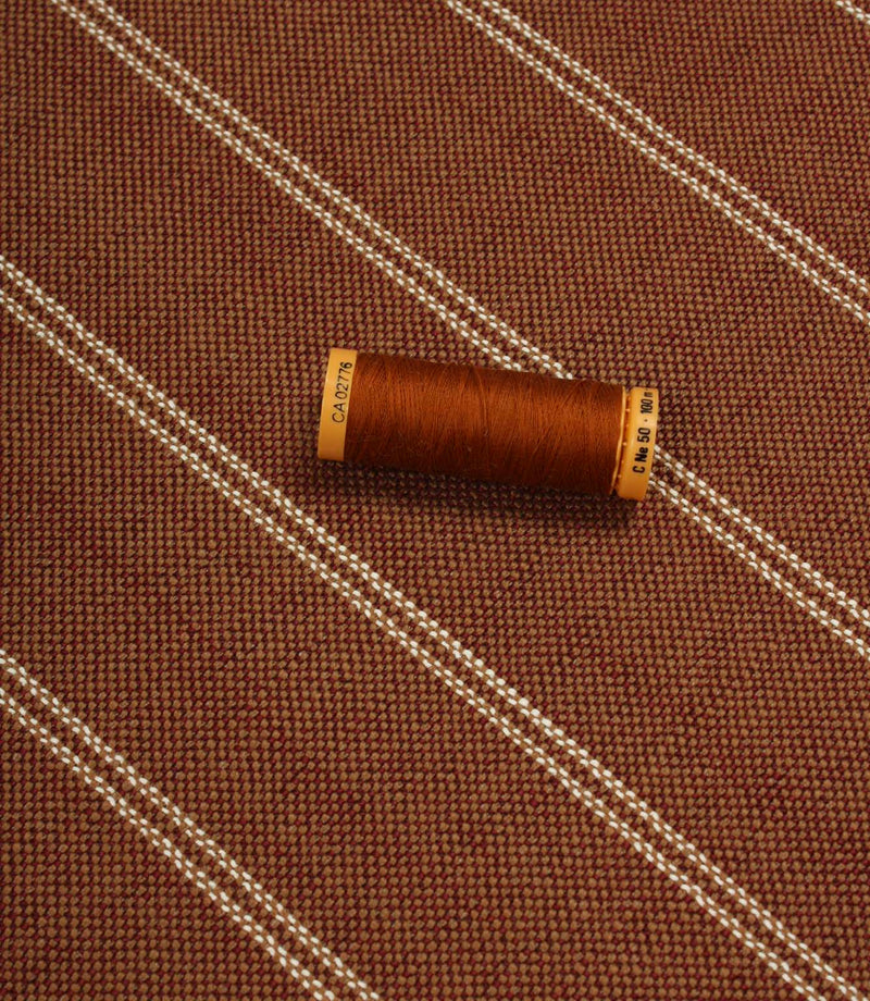 Copper striped upholstery fabric displayed with a matching thread spool to highlight scale and woven texture.