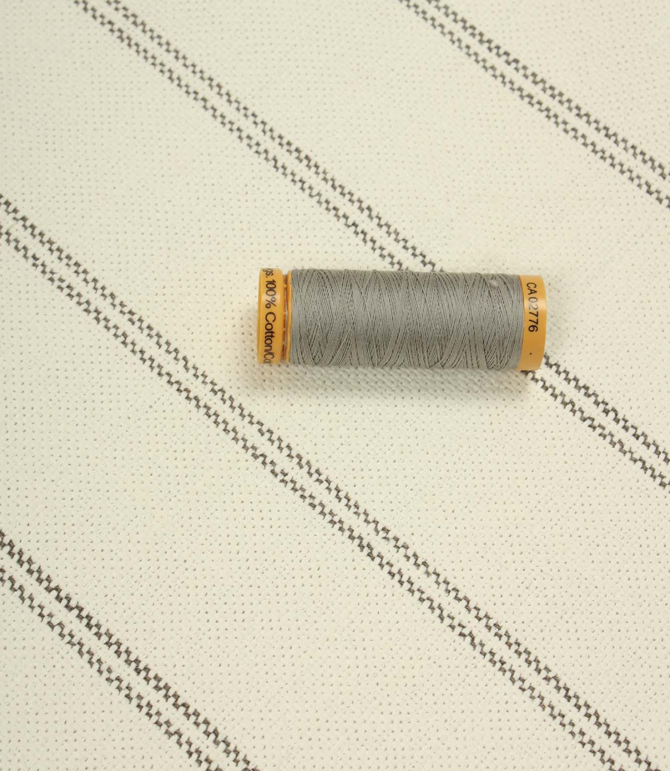 Ecru striped upholstery fabric displayed with a grey thread spool to highlight scale and woven texture.