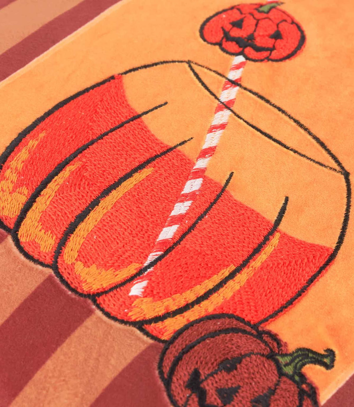 Close-up of the Pumpkin Spice velvet cushion showing the orange and red embroidered pumpkin-shaped drink, striped straw and jack-o'-lantern details on soft velvet.
