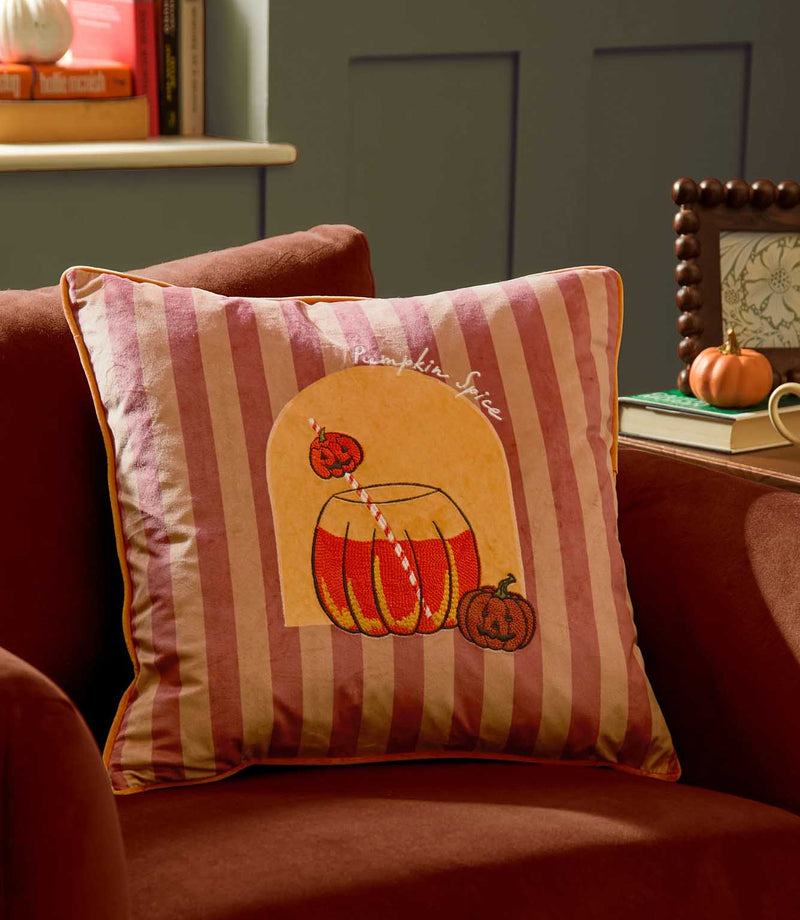 Pumpkin Spice velvet cushion styled on a rust orange armchair, featuring pink and burgundy stripes, orange piping and an embroidered pumpkin drink design in an autumn interior.