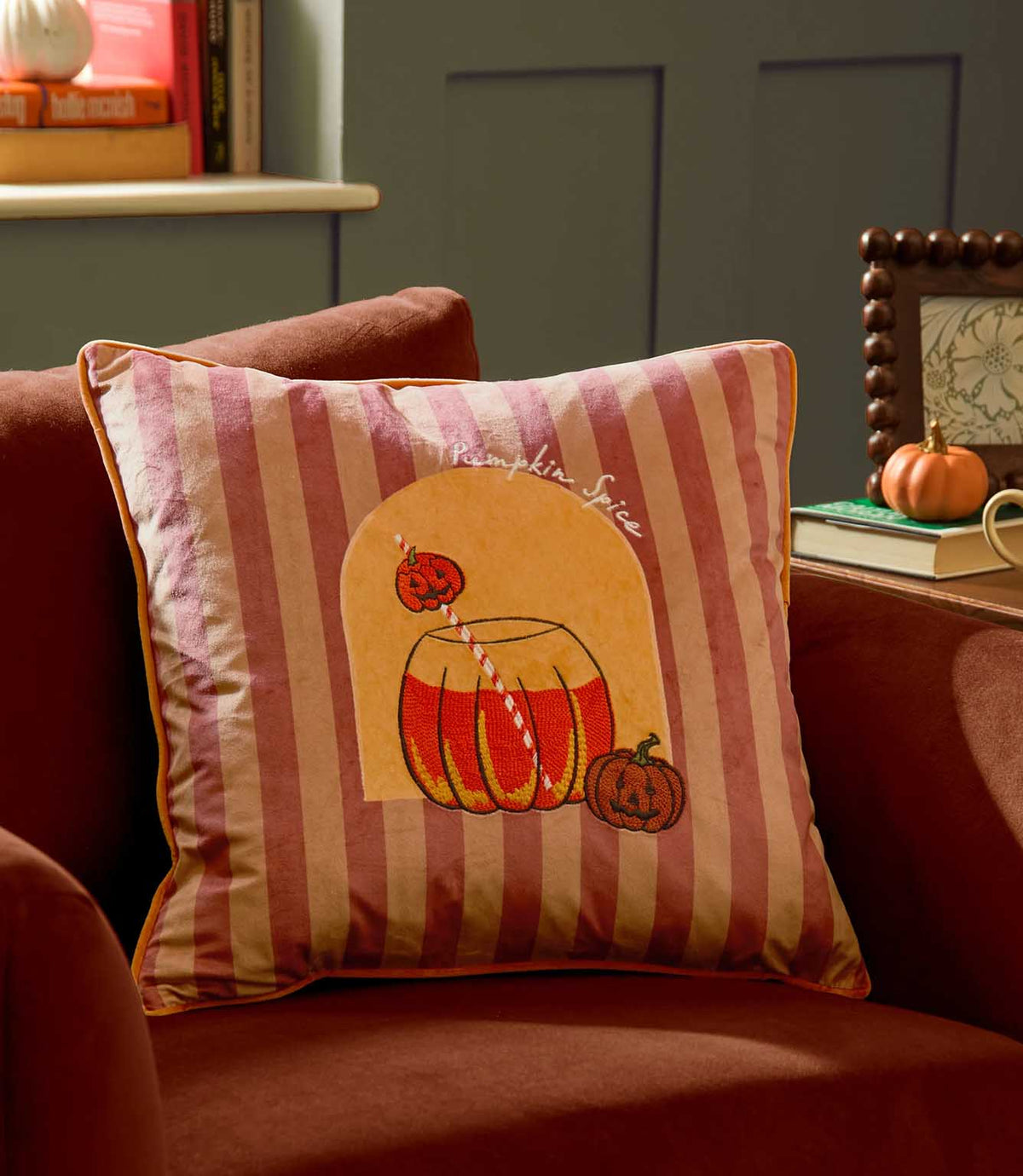 Pumpkin Spice velvet cushion styled on a rust orange armchair, featuring pink and burgundy stripes, orange piping and an embroidered pumpkin drink design in an autumn interior.
