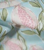 Macro photograph of Rithani Robin's Egg fabric highlighting the fine woven texture and detailed pomegranate illustration, with pink patterned seeds, cream fruit outlines and sage green leaves on a pale blue background.