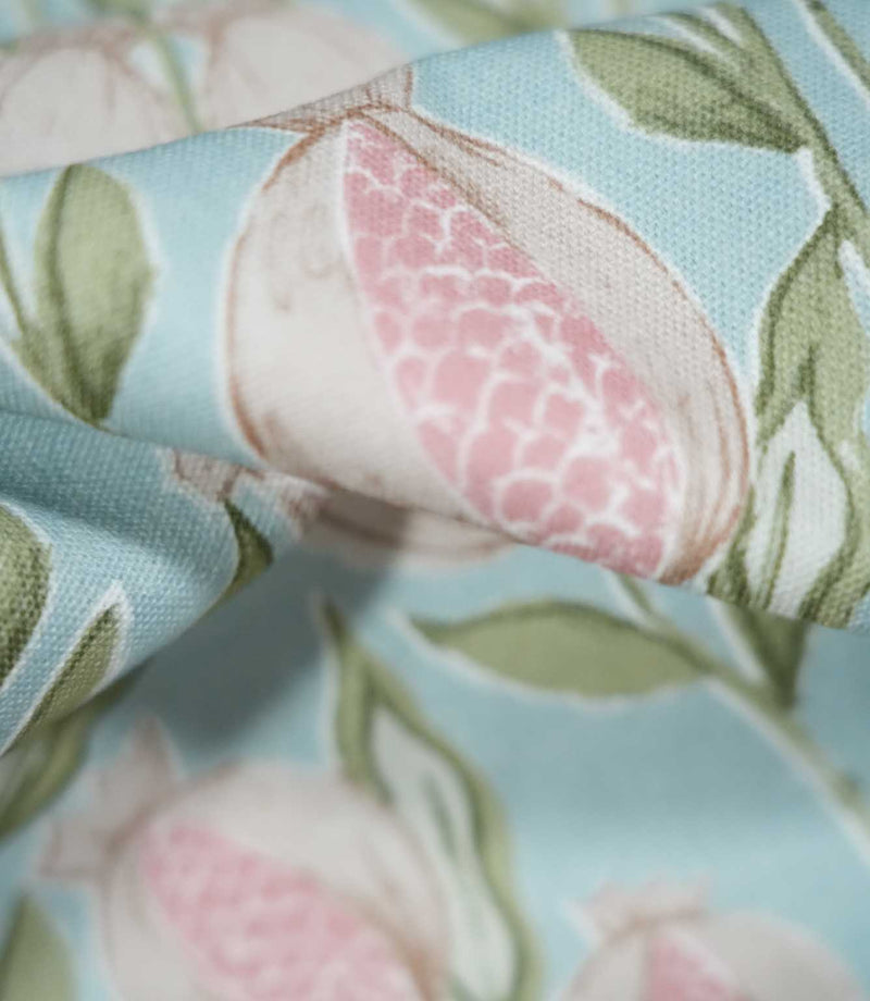 Macro photograph of Rithani Robin's Egg fabric highlighting the fine woven texture and detailed pomegranate illustration, with pink patterned seeds, cream fruit outlines and sage green leaves on a pale blue background.