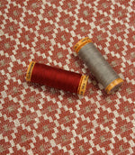 Geometric woven furnishing fabric displayed with coordinating thread reels, featuring a warm rust red and cream patterned design.