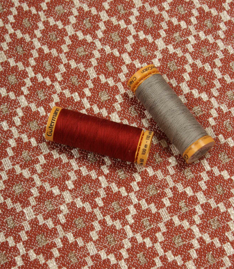 Geometric woven furnishing fabric displayed with coordinating thread reels, featuring a warm rust red and cream patterned design.
