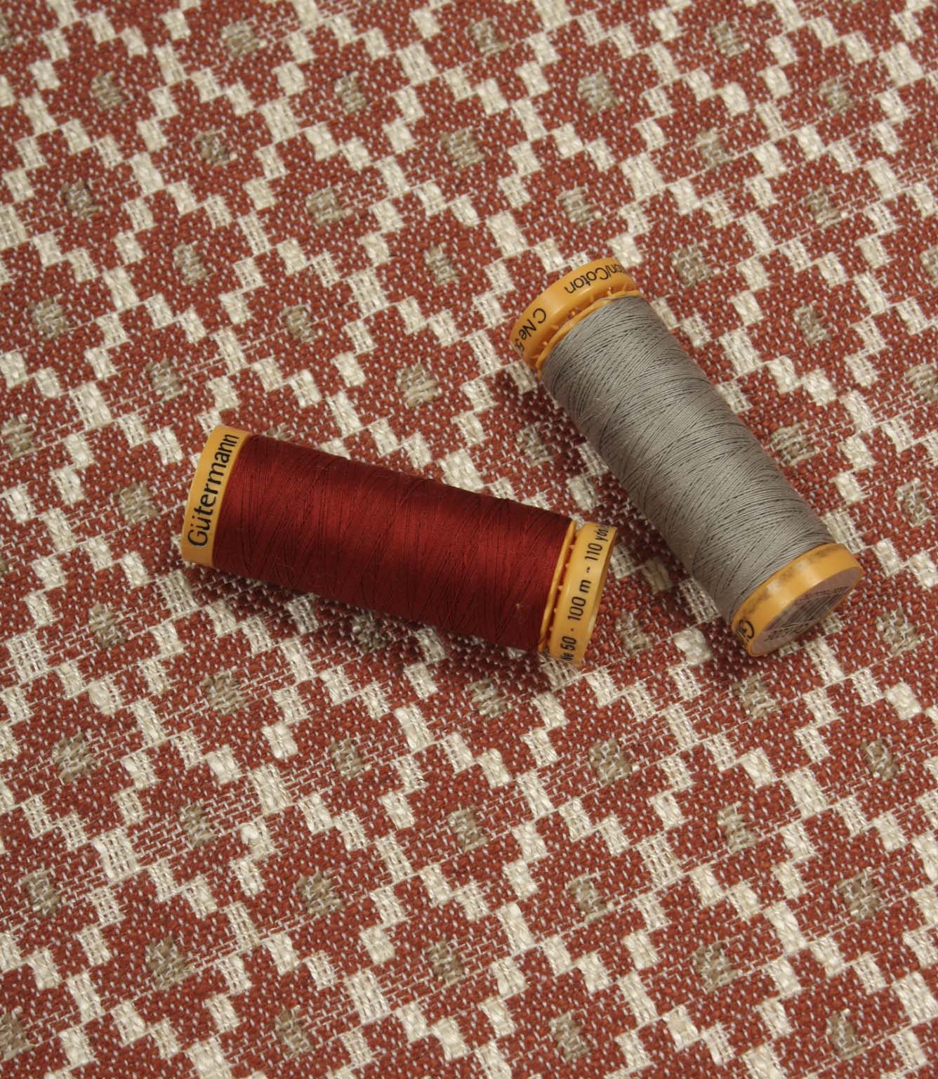 Geometric woven furnishing fabric displayed with coordinating thread reels, featuring a warm rust red and cream patterned design.