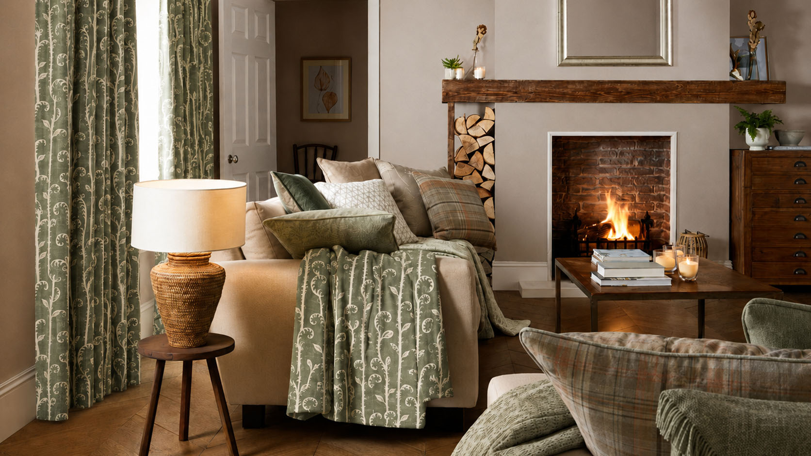 A cosy living room scene featuring sage green patterned curtains, a matching throw blanket, and pillows on a beige sofa, with a lit fireplace in the background.